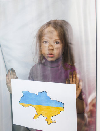 a little Ukrainian girl with sad eyes stands behind the glass, holding a drawn map of Ukraine in her hands. children and warの写真素材