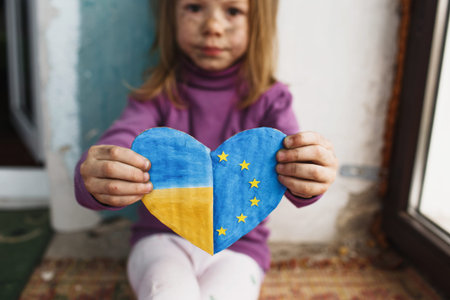 a little Ukrainian girl with a face blackened by fire is sitting on the floor by the window holding...