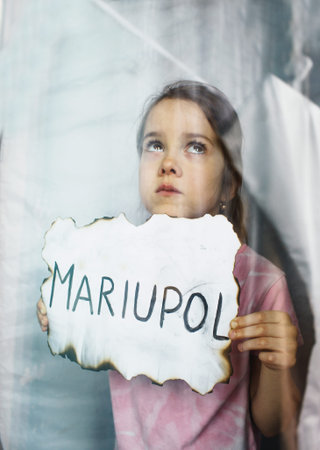 a little Ukrainian girl with sad eyes is standing behind the glass holding a piece of burnt paper...