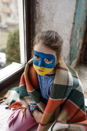 a little girl with a Ukrainian flag painted on her face is sitting covered with a blanket by the window. Children and the war in Ukraineの写真素材