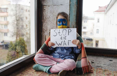 a little girl with a Ukrainian flag painted on her face is sitting under a blanket with a Stop the War poster. children and warの写真素材