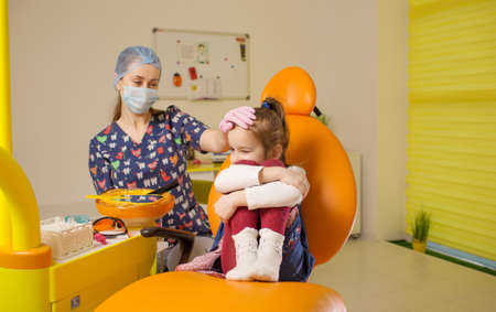 a girl dentist calms a frustrated little girl sitting in a dental chair at a doctor's appointmentの写真素材