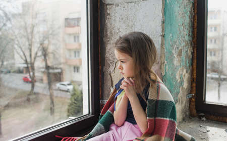 a little Ukrainian girl prays for the freedom of Ukraine holding ribbons in the color of the flag. Sitting by the window and sad.の写真素材