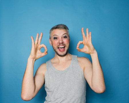 An LGBT gay man smiles on a blue background and shows the excellent sign.