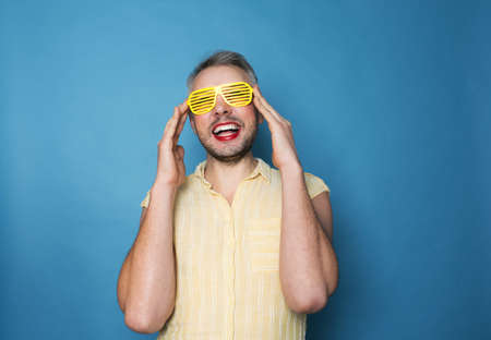 An Lgbt transsexual man with makeup on his face in yellow stylish glasses smiles on a blue isolated background.の写真素材