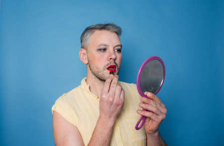 an Lgbt man with makeup on his face paints his lips looking in the mirror on a blue isolated background.の写真素材