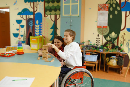 a girl in a wheelchair is playing board games sitting at a table. A female psychologist is engaged...