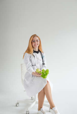 An attractive female doctor in a white coat is sitting smiling and holding an apple and salad in her hands. A cheerful nutritionist who recommends a healthy diet to the patient.の写真素材