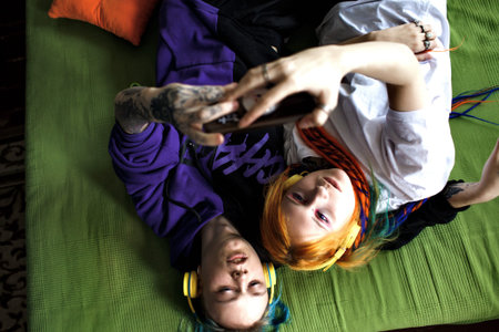 a stylish young modern couple with multicolored hair are lying on the bed looking at the smartphone...
