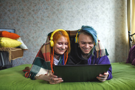 Portrait of a young tattooed punk couple of a girl and a guy with long dyed hair braided, lying on a large green bed. They listen to music. Yellow large headphones. smartphone. Stylish modern youthの写真素材