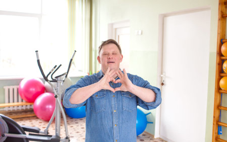 A young man with Down syndrome, dressed in a casual denim jacket smiling in the shape of a heart...