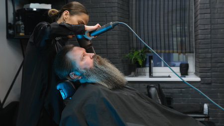 a hairdresser girl using a hair dryer to straighten a man's long beard in a barbershopの写真素材