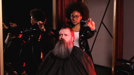 an American hairdresser girl holds scissors in her hands, looks at the camera and smiles. A man with a big beard is sitting in a barber chairの写真素材