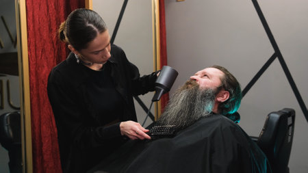 a hairdresser girl using a hair dryer to dry a man's long beard in a barbershop.の写真素材