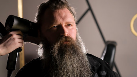 a young stylist does styling with a hair dryer to a bearded man sitting in a barber chairの写真素材