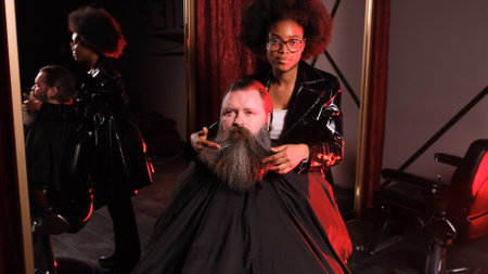 an African-American hairdresser girl straightens her beard with her hands to a male client sitting in an armchairの写真素材