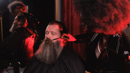 An African-American stylist girl in a black leather raincoat combs the beard of a client in a barber shopの写真素材