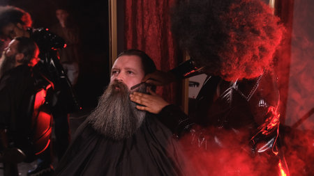 An African-American stylist girl in a black leather raincoat combs the beard of a client in a barber shopの写真素材