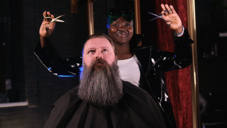 an American hairdresser girl holds scissors in her hands, looks at the camera and smiles. A man with a big beard is sitting in a barber chairの写真素材