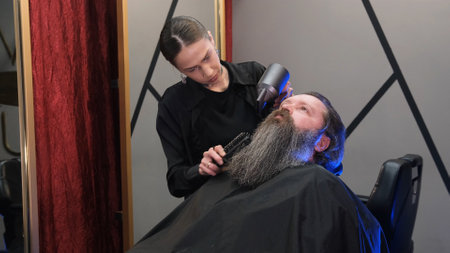 a hairdresser girl using a hair dryer to dry a man's long beard in a barbershop.の写真素材