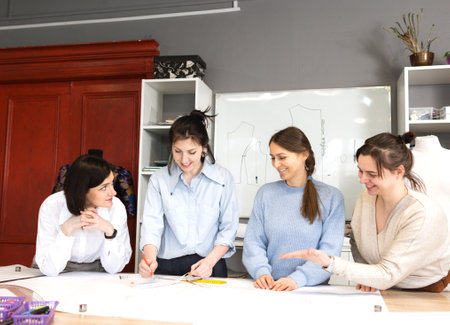 Four female fashion designers work together in a tailoring studio. A group of women dressmakers discusses fabric, pattern and sketch in a sewing workshop. The concept of small businessの写真素材