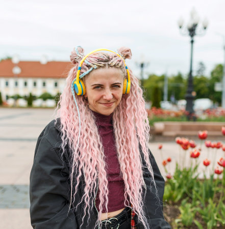 A young beautiful girl with pink hair listens to music with headphones on a city street.の写真素材
