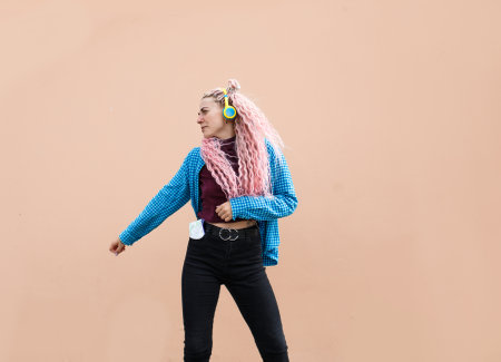 A happy young woman with pink long dreadlocked hair and a blue shirt is dancing to music with headphones on a beige background. Empty space for advertising text.の写真素材