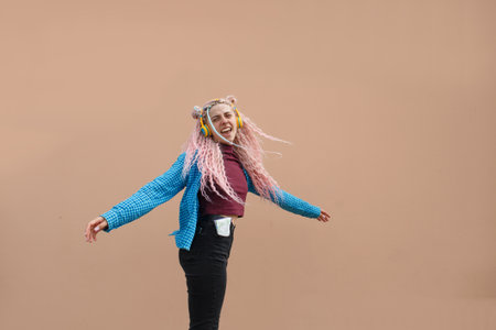 A happy young woman with pink long dreadlocked hair and a blue shirt is dancing to music with headphones on a beige background. Empty space for advertising text.の写真素材