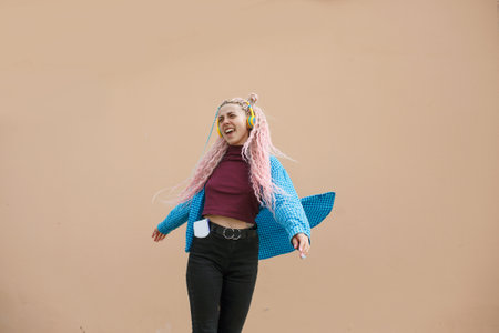 A happy young woman with pink long dreadlocked hair and a blue shirt is dancing to music with headphones on a beige background. Empty space for advertising text.の写真素材