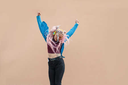 A happy young woman with pink long dreadlocked hair and a blue shirt, rhythmically dancing to music with headphones on a beige background. Empty space for advertising text.の写真素材