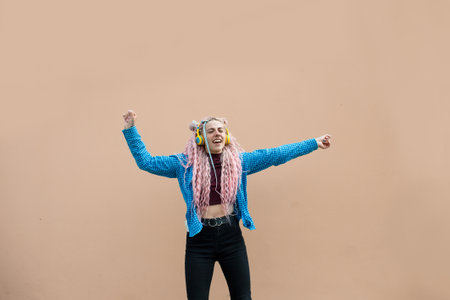 An attractive young woman with pink long dreadlocked hair and a blue shirt, rhythmically dancing to music with headphones on a beige background. Empty space for advertising text.の写真素材