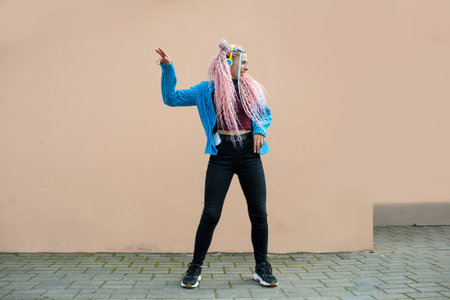 An attractive young woman with pink long dreadlocked hair and a blue shirt, rhythmically dancing to music with headphones on a beige background. Empty space for advertising text.の写真素材