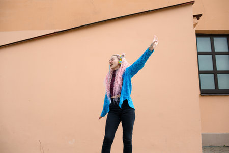 An attractive young woman with pink long dreadlocked hair and a blue shirt, rhythmically dancing to music with headphones on a beige background. Empty space for advertising text.の写真素材