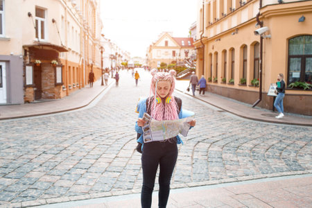 An attractive young tourist in a blue shirt is exploring a new city. A girl with long pink hair holds a paper map of the city scheme in her hands. Travel around Europeの写真素材