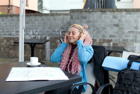 An attractive young tourist in a blue shirt drinks coffee and listens to music with headphones. A girl with long pink hair is having lunch in a cafe. Travel around Europeの写真素材
