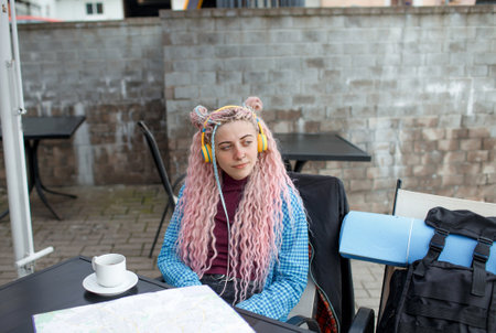 An attractive young tourist in a blue shirt drinks coffee and listens to music with headphones. A girl with long pink hair is having lunch in a cafe. Travel around Europeの写真素材