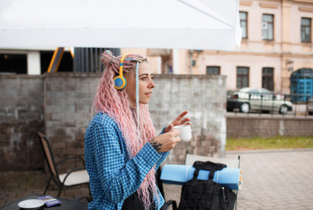 A young tourist girl in a blue shirt drinks coffee and listens to music with headphones. A girl with long pink hair is having lunch in a cafe. Travel around Europeの写真素材
