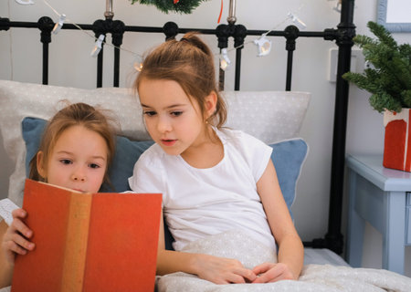 Two little sisters are reading a book in a bed decorated with Christmas lightsの写真素材