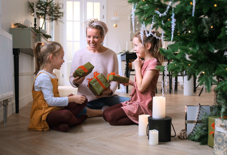 Cheerful mom and her cute daughters exchange Christmas gifts sitting on the floor near the Christmas tree.の写真素材