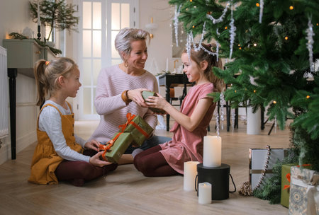 Cheerful mom and her cute daughters exchange Christmas gifts sitting on the floor near the Christmas tree.の写真素材