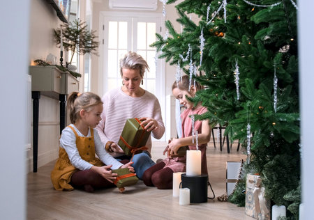 Cheerful mom and her cute daughters exchange Christmas gifts sitting on the floor near the Christmas tree.の写真素材
