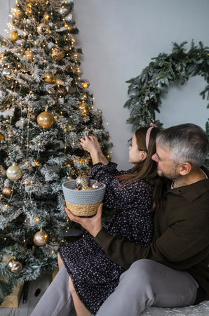 A father and daughter decorate their Christmas tree together. The girl adds ornaments to the flocked tree, while her dad holds a basket of decorations. Warm lighting creates a cozy, festive atmosphere in their home.の写真素材