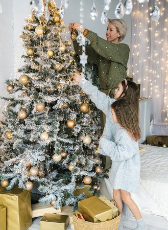 A mother and her two daughters create magical Christmas memories while decorating a snow-flocked tree with elegant gold ornaments. The warm lighting and festive atmosphere radiate joy and family love.の写真素材