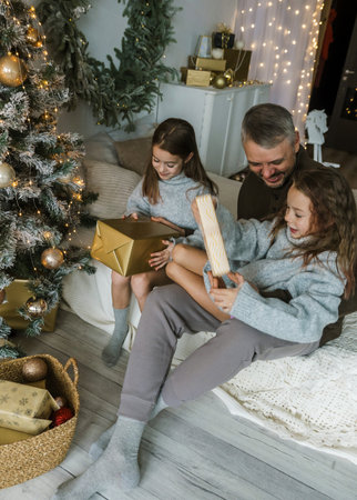 A father and two daughters share the joy of Christmas morning, opening presents on a bed. The festive atmosphere is filled with warmth and laughter.の写真素材
