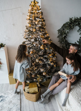 A heartwarming Christmas scene: A father helps his two daughters decorate a beautifully lit Christmas tree. The family's joy and festive spirit are contagious.の写真素材
