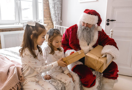 A jolly Santa Claus is seated on a bed with two little girls in matching pajamas. He is holding a wrapped gift while the girls look excitedly at him. There is a second gift on the bed, suggesting Santa is about to give them their presents.の写真素材