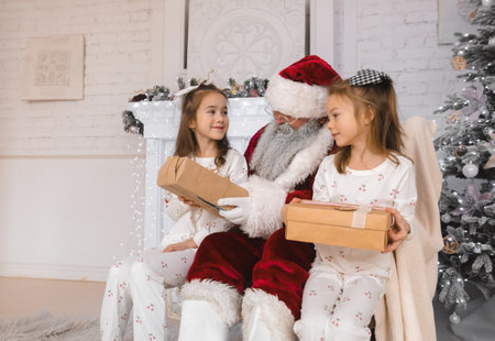 Santa Claus sits on a white fur rug next to two young girls, who are opening presents. They are dressed in white pajamas with red cherries on them. Santa is wearing a red suit and a white beard, and he is holding a gift. The girls are looking at their presents and smiling.の写真素材