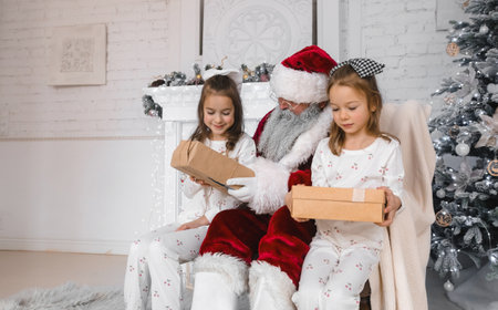 Santa Claus shares the Christmas magic with two adorable girls in matching pajamas, each receiving a thoughtfully wrapped gift. The heartwarming scene is set against a backdrop of a festively decorated fireplace and Christmas tree, creating a cozy and enchanting holiday atmosphere.の写真素材