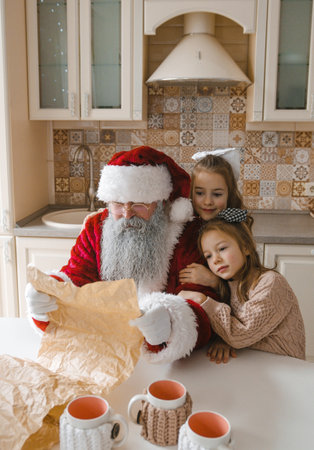 Santa Claus reads Christmas wishes from two sweet girls huddled close in a cozy kitchen. A heartwarming scene perfect for holiday cards, websites, and social media. Christmas magicの写真素材