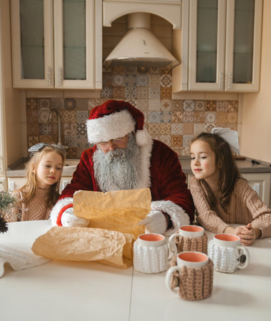 Santa Claus reads a Christmas wish list with two adorable girls in a warm, inviting kitchen. Cozy scene perfect for holiday cards and family-themed projects.の写真素材
